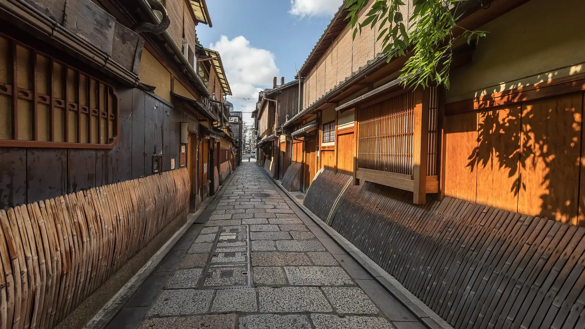 A stone-paved lane in Gion, Kyoto, between wooden townhouses with bamboo fences and slatted windows, in warm sunlight